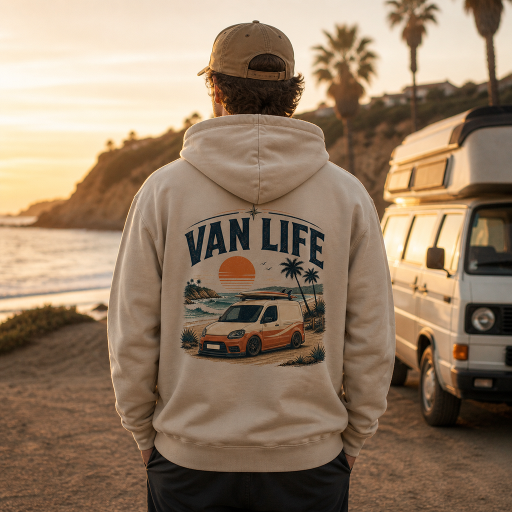 Person wearing a 'Van Life' hoodie with a sunset and van design, standing on a beach with a van in the background.