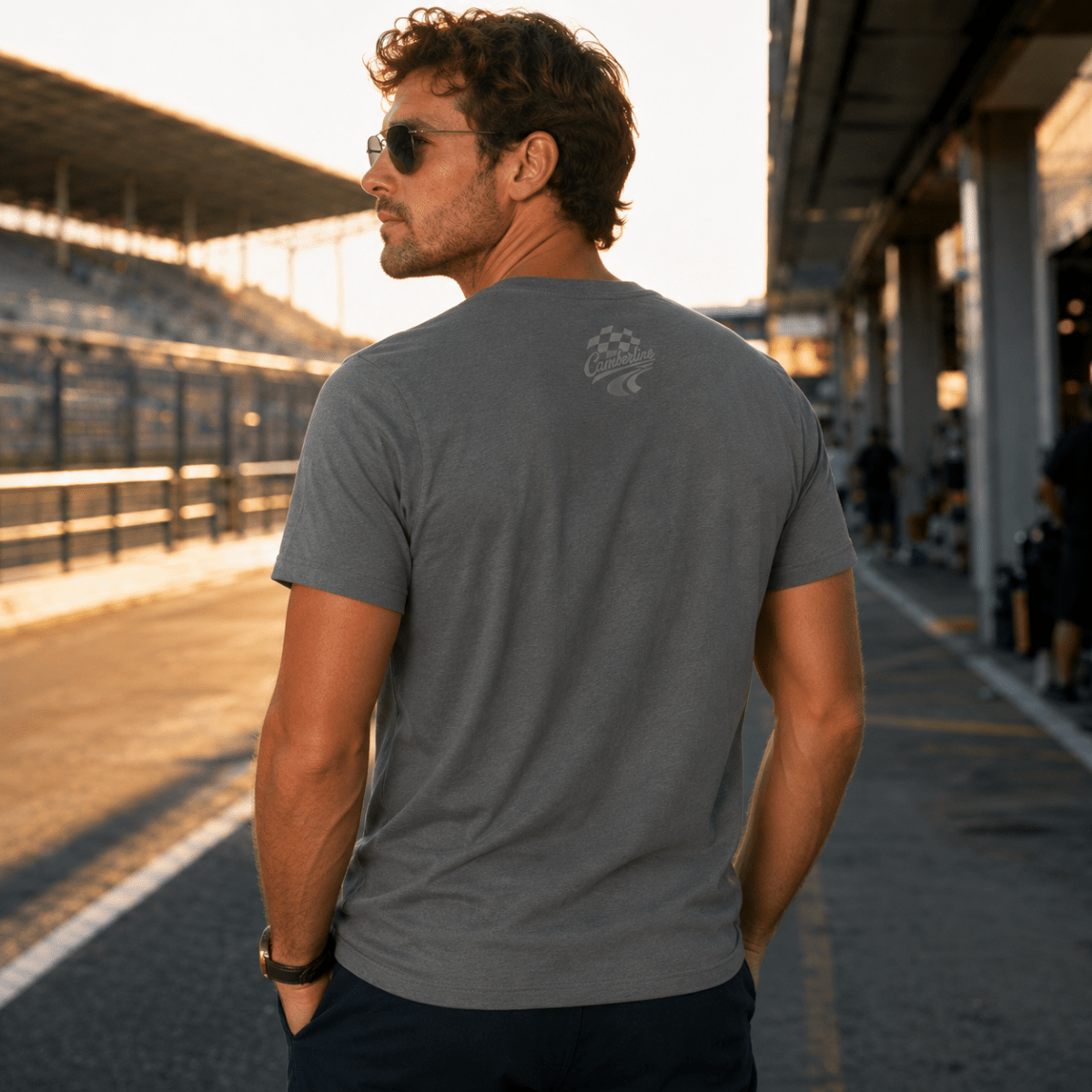 A man wearing a dark gray crewneck t-shirt with a small white crest logo on the upper back, standing on a racetrack at sunset.