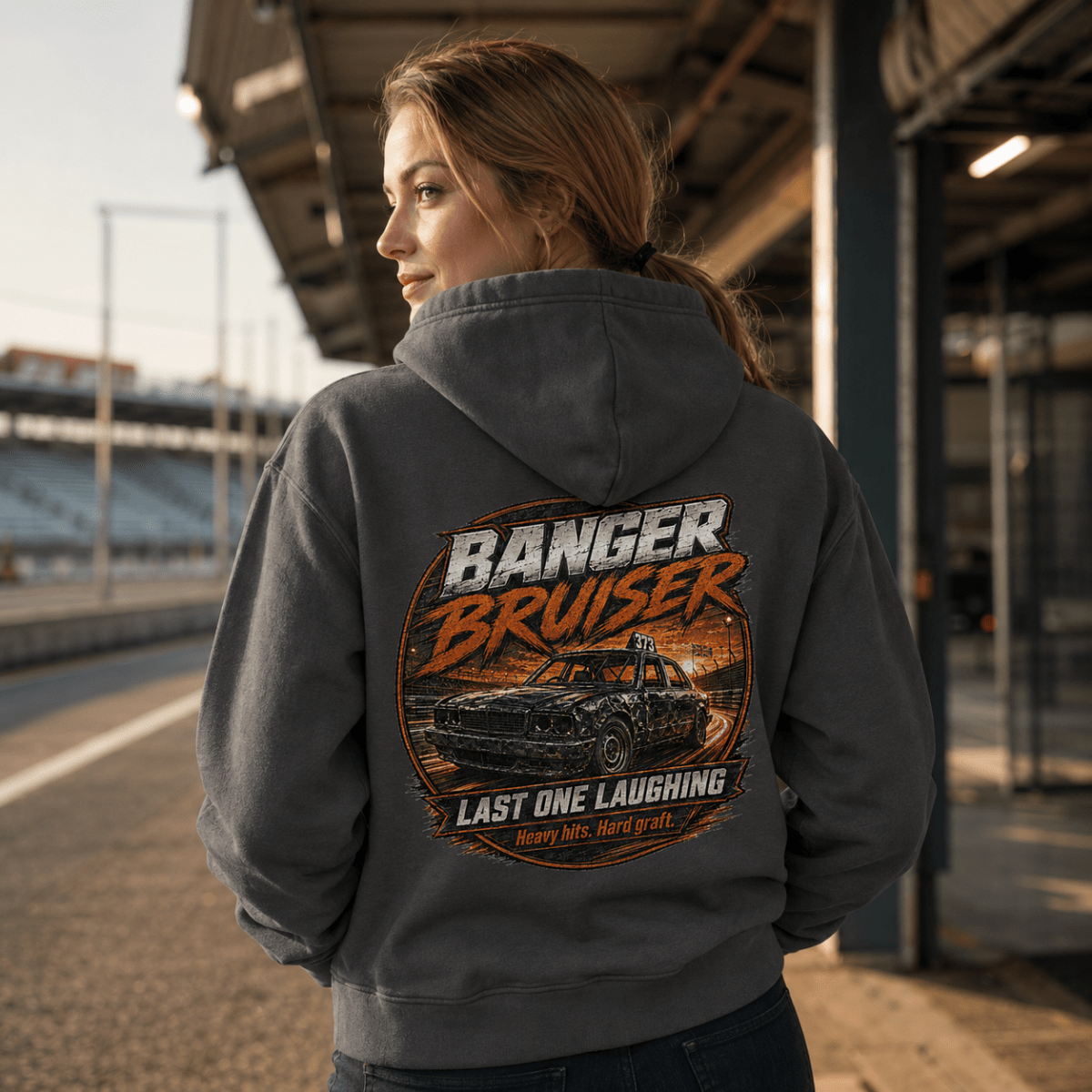 A woman stands outdoors on a racetrack wearing a dark gray hoodie from the back, which features a large graphic print of a black car and the text 'BANGER BRUISER' and 'LAST ONE LAUGHING'.