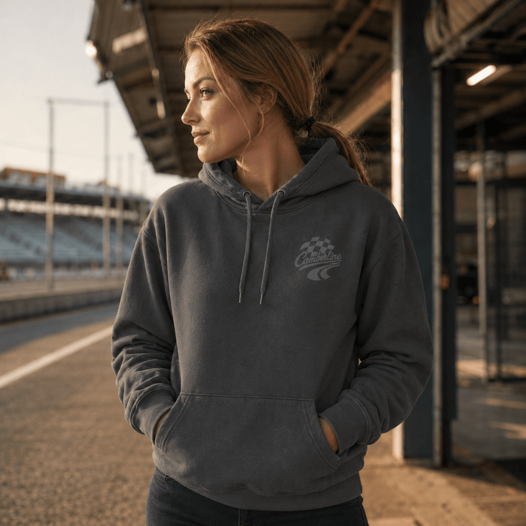A woman wearing a dark gray hooded sweatshirt with a small racing-style chest graphic, standing on a racetrack under a covered area at sunset.