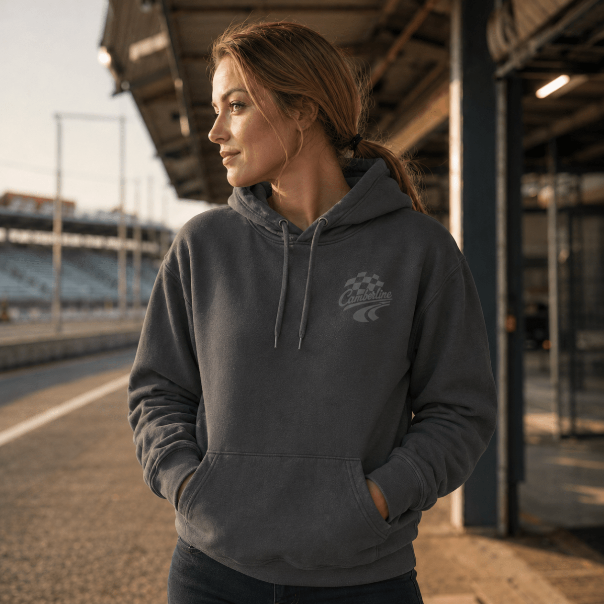 A woman wearing a dark gray hooded sweatshirt with a small racing-style chest graphic, standing on a racetrack under a covered area at sunset.