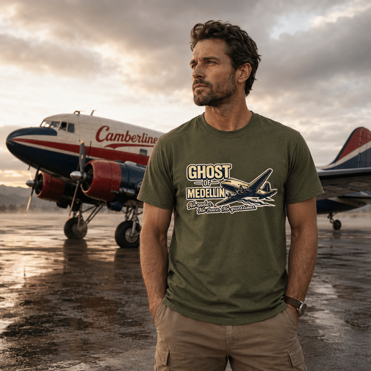 Man wearing a green t-shirt with an airplane graphic and text, standing in front of vintage airplanes on a runway.