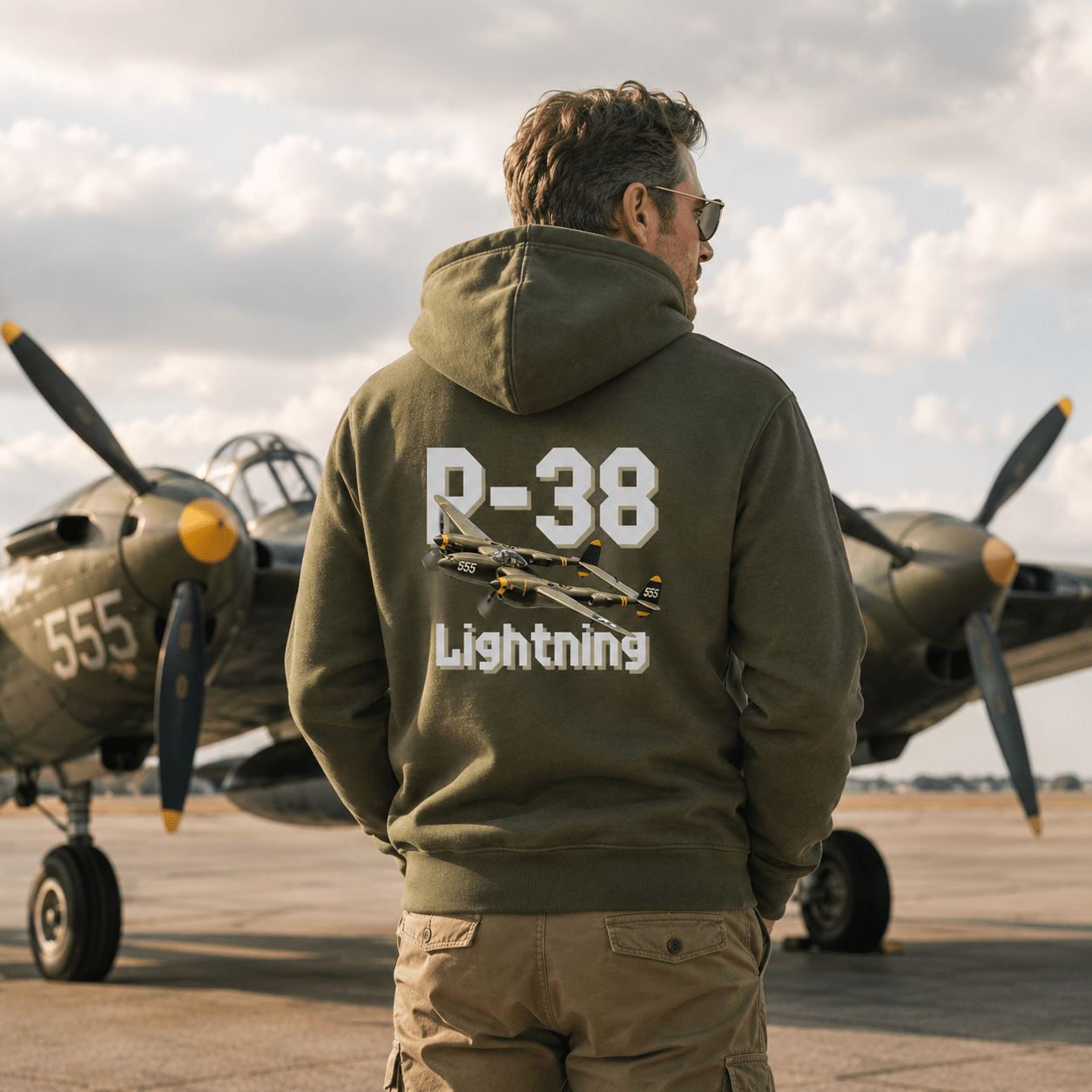 Man wearing a green hoodie with 'P-38 Lightning' design in front of vintage airplanes.