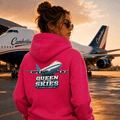 Person wearing a pink hoodie with an airplane design and 'Queen of the Skies' text, standing in front of a Camberline airplane.