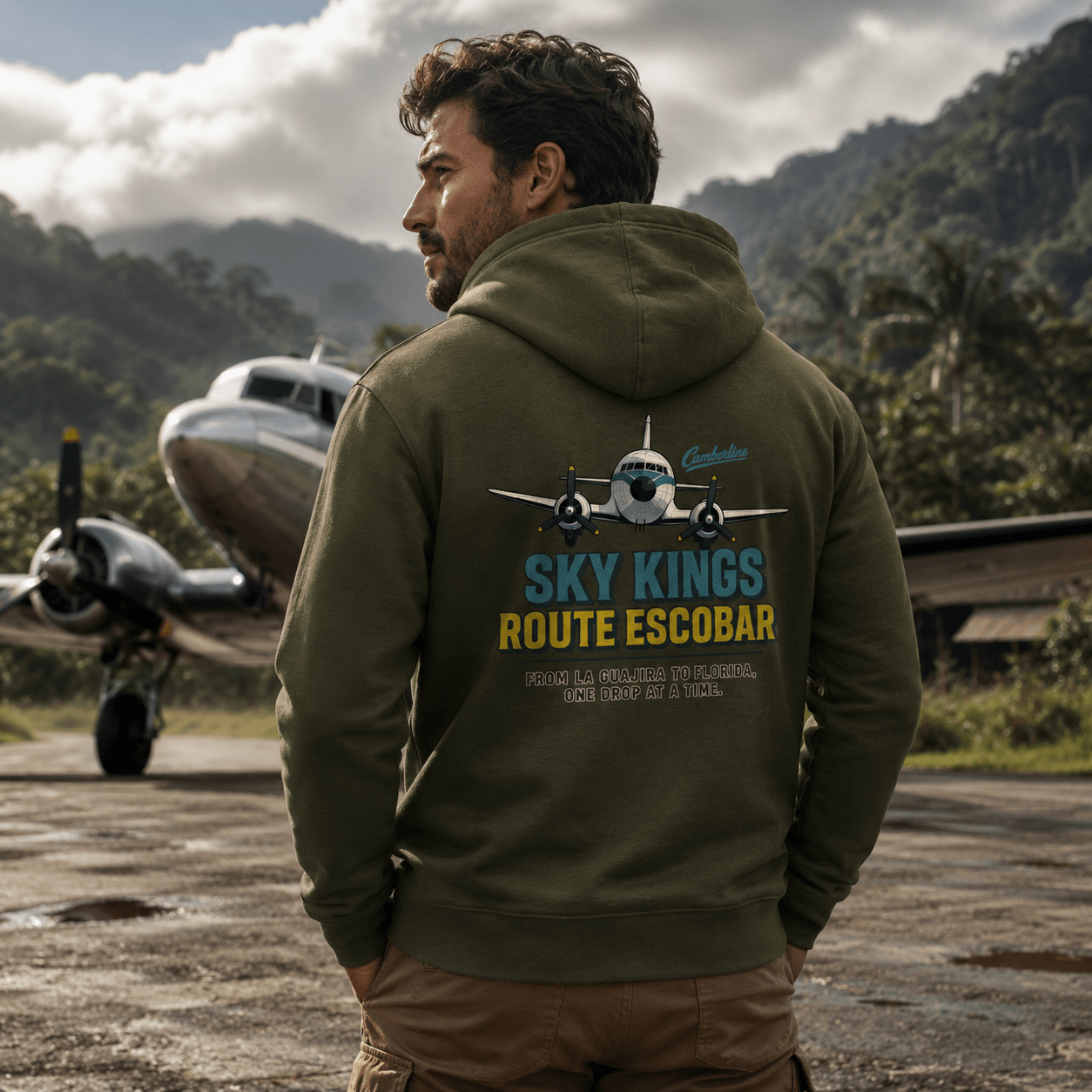 Man wearing a hoodie with 'Sky Kings Route Escobar' design, standing on an airstrip with an airplane in the background.