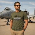 Man wearing a green t-shirt with 'Warthog' design in front of an airplane on a runway.