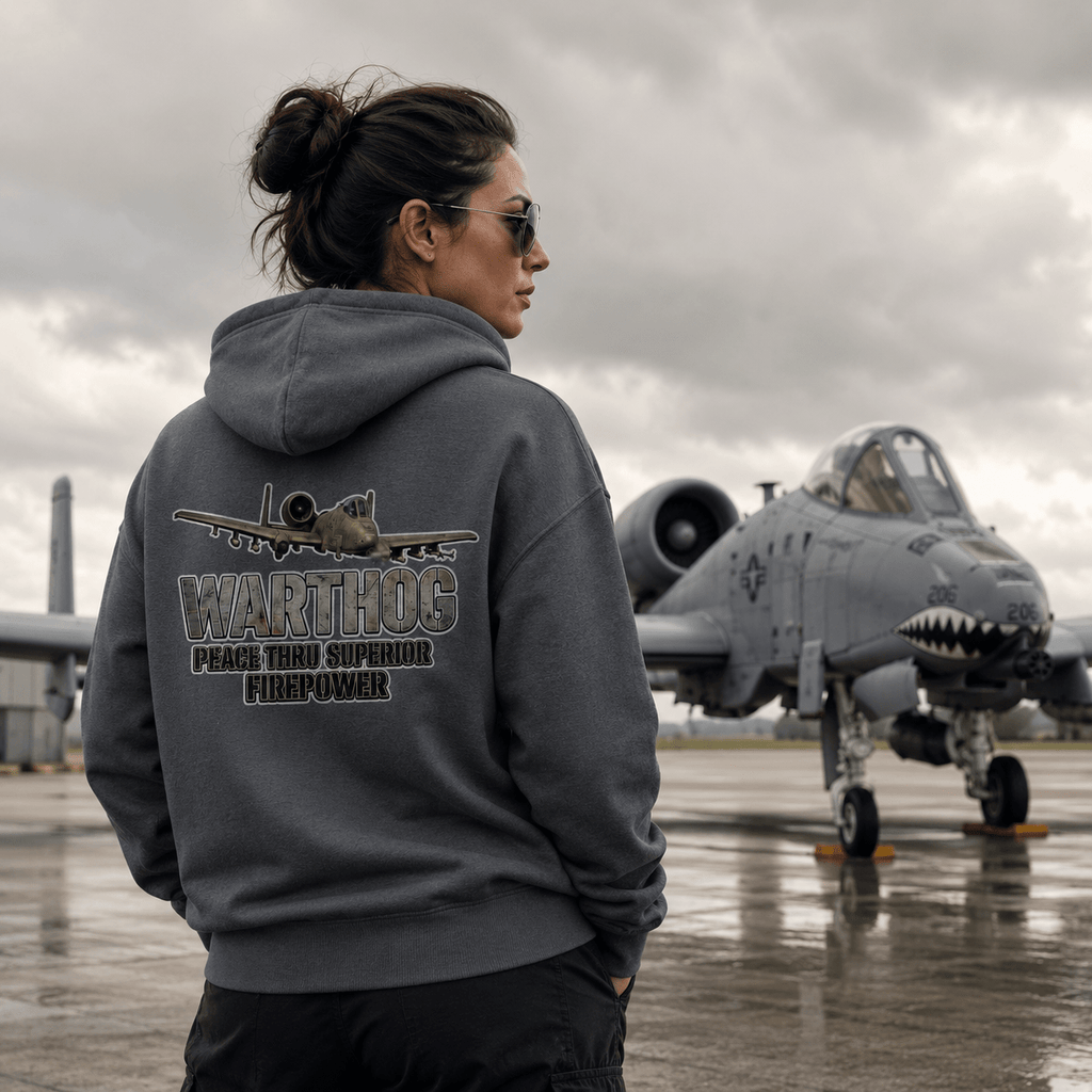 Person wearing a hoodie with an airplane design and text, standing in front of a military aircraft on a runway.