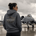 Person wearing a hoodie with an airplane design and text, standing in front of a military aircraft on a runway.