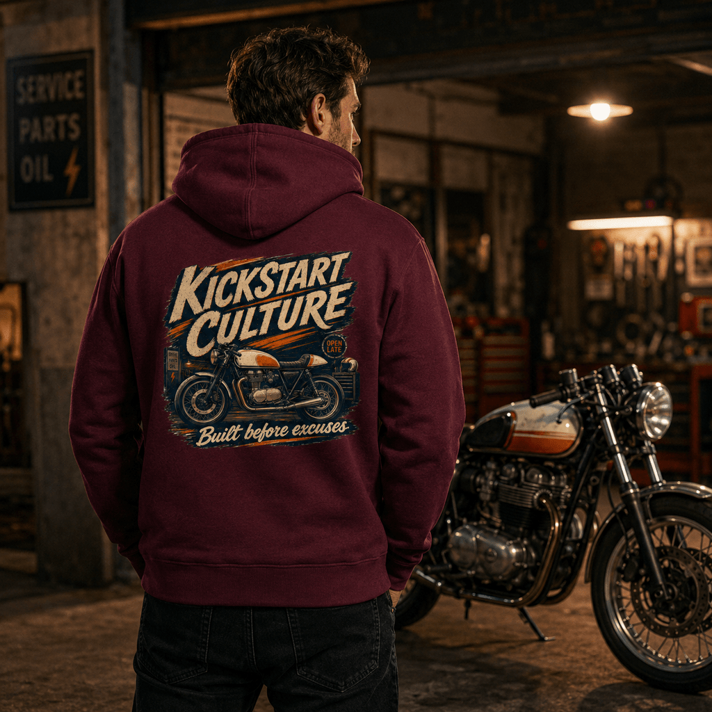 Man wearing burgundy Kickstart Culture café racer motorcycle hoodie showing the back graphic, standing in a vintage garage next to a classic café racer motorcycle