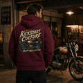Man wearing burgundy Kickstart Culture café racer motorcycle hoodie showing the back graphic, standing in a vintage garage next to a classic café racer motorcycle