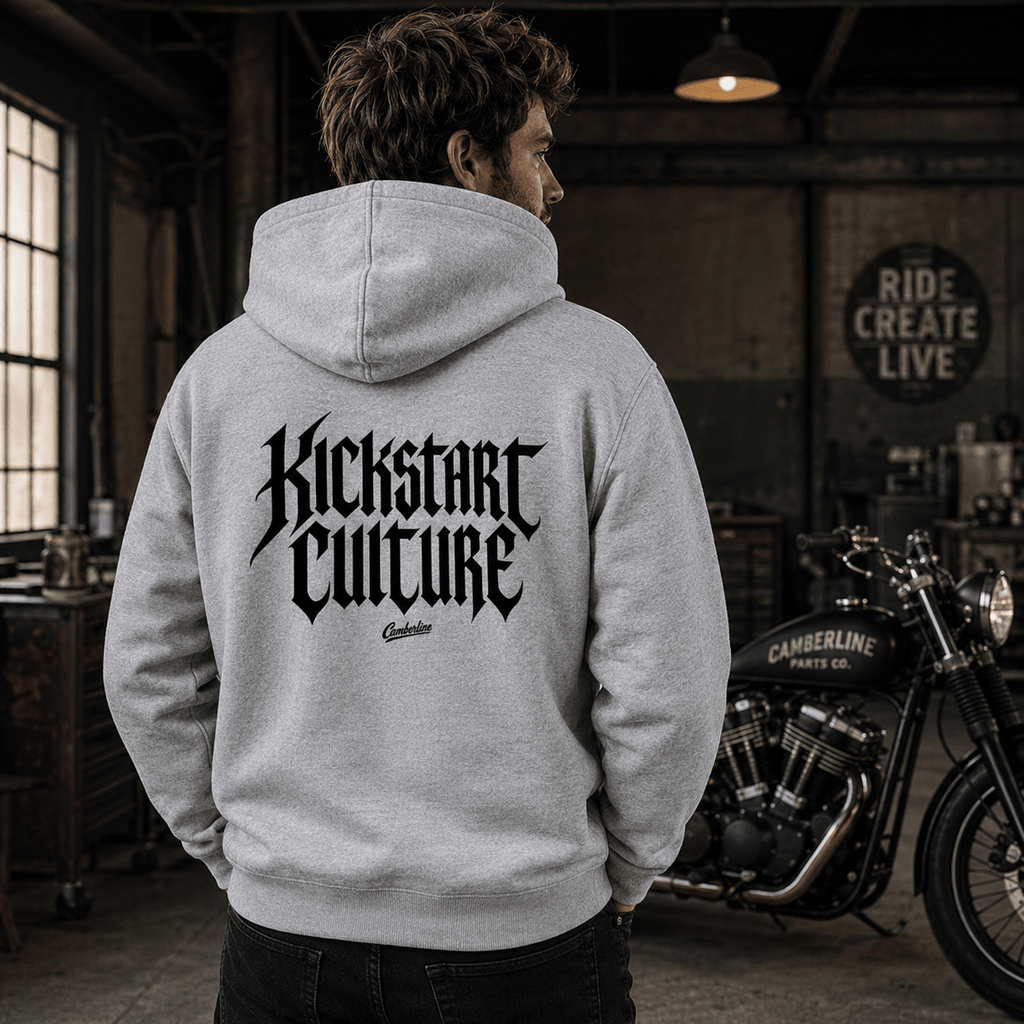 Man wearing a heather grey Kickstart Culture gothic typography motorcycle hoodie showing the bold blackletter back print, standing in a vintage motorcycle workshop with a Camberline Parts Co. motorcycle