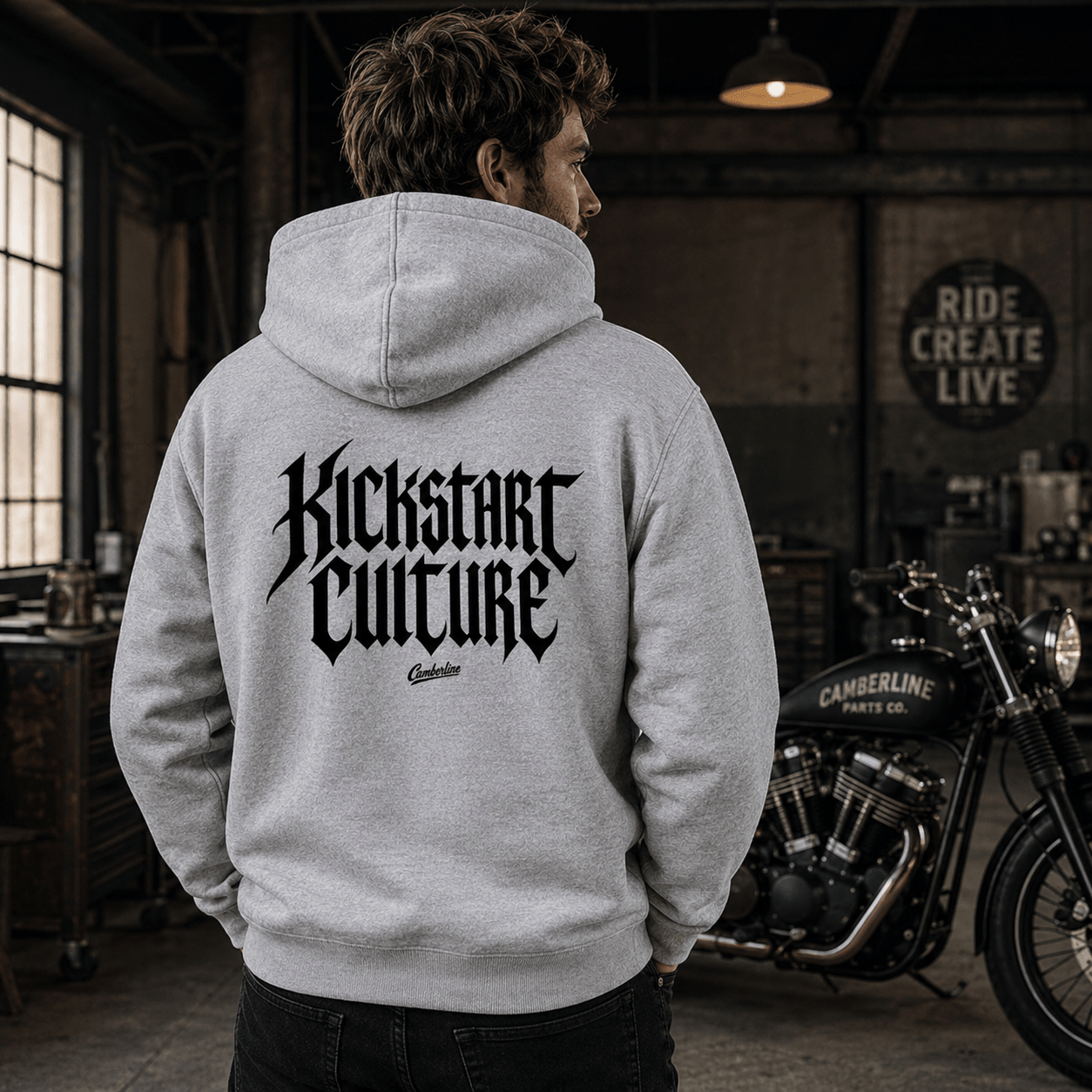 Man wearing a heather grey Kickstart Culture gothic typography motorcycle hoodie showing the bold blackletter back print, standing in a vintage motorcycle workshop with a Camberline Parts Co. motorcycle