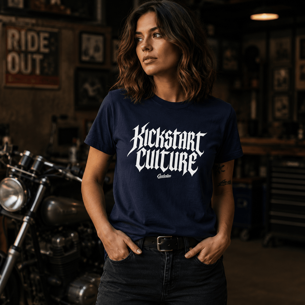 Woman wearing a navy Kickstart Culture gothic typography motorcycle t-shirt with bold white blackletter front print, standing in a vintage motorcycle garage