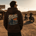 Man wearing a hot chocolate brown Ride Out Syndicate motorcycle hoodie showing the vintage pack rider back graphic, standing on a desert highway at golden hour with motorcyclists approaching in the background