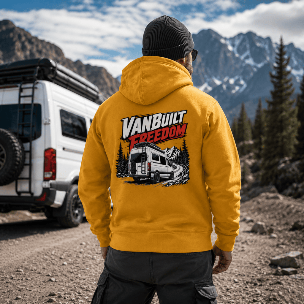 Model wearing Camberline amber/mustard van life hoodie showing VanBuilt Freedom rear-view high-roof van with ladder on mountain road on back