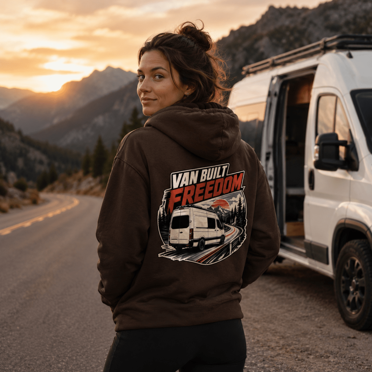 Model wearing Camberline chocolate brown van life hoodie showing Van Built Freedom panel van on elevated road at sunset on back