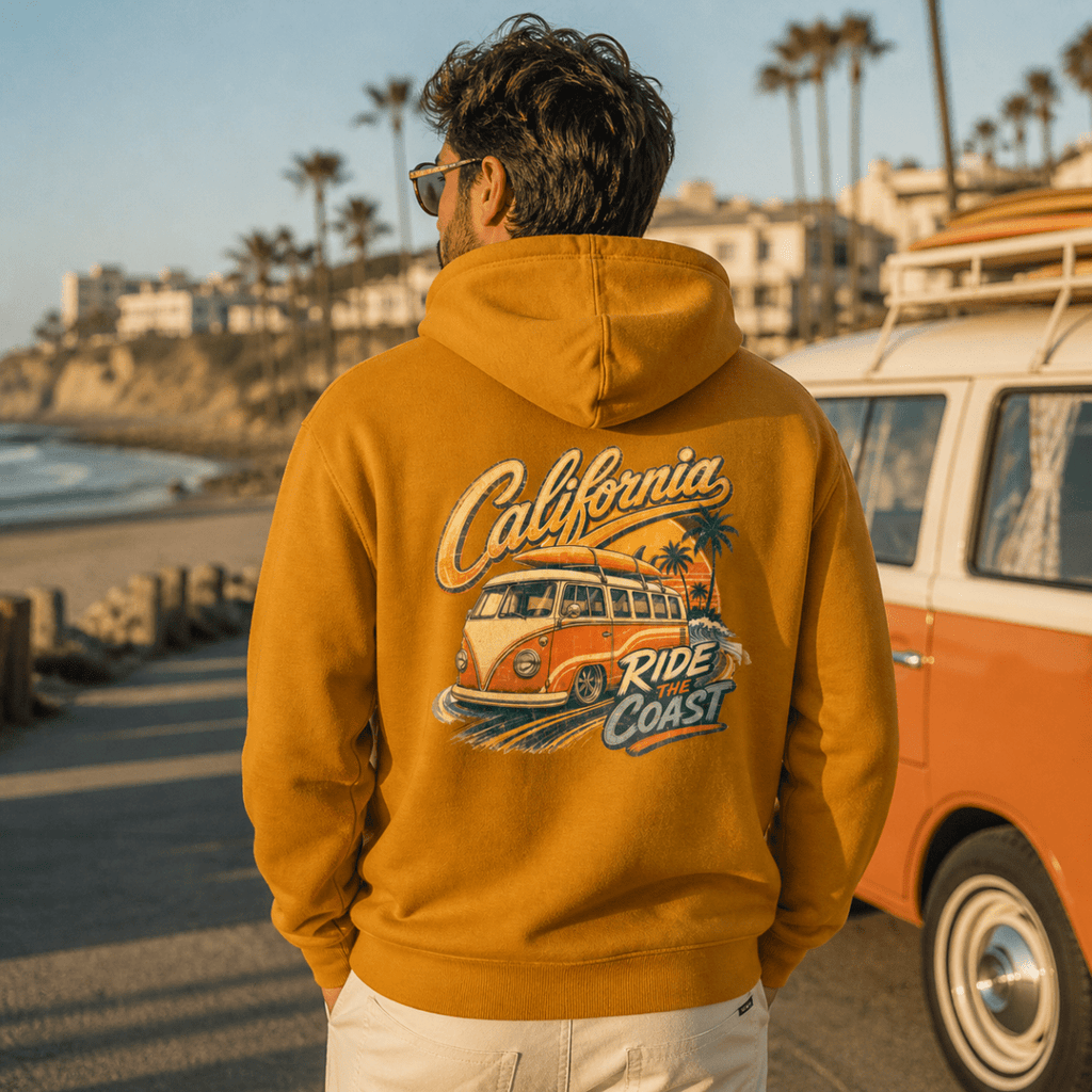 Person wearing a mustard yellow hoodie with 'California Ride the Coast' design, standing near a vintage van by a beach.