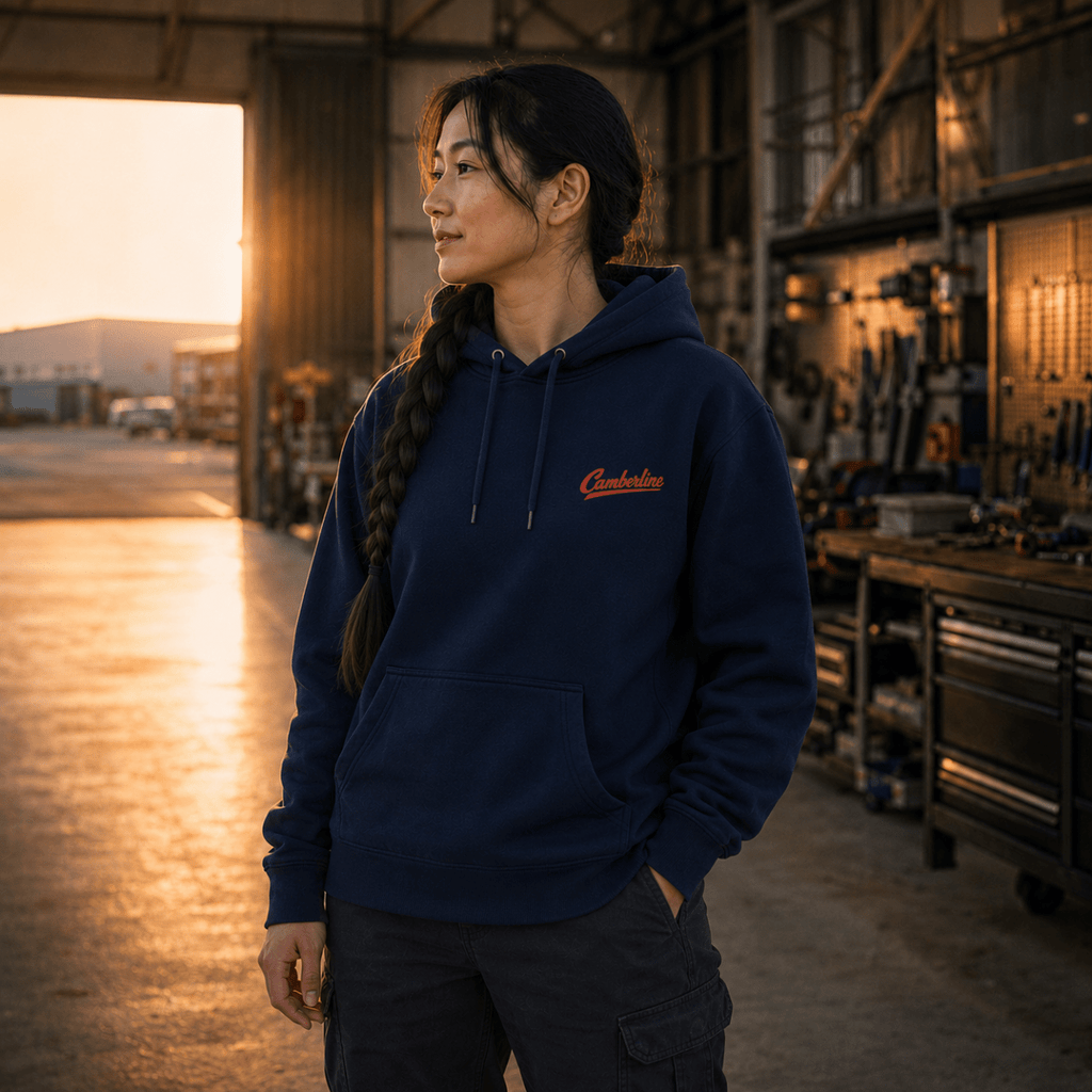 A woman wearing a navy blue hoodie with a red embroidered 'Carmen' logo on the chest, standing in a workshop during golden hour.
