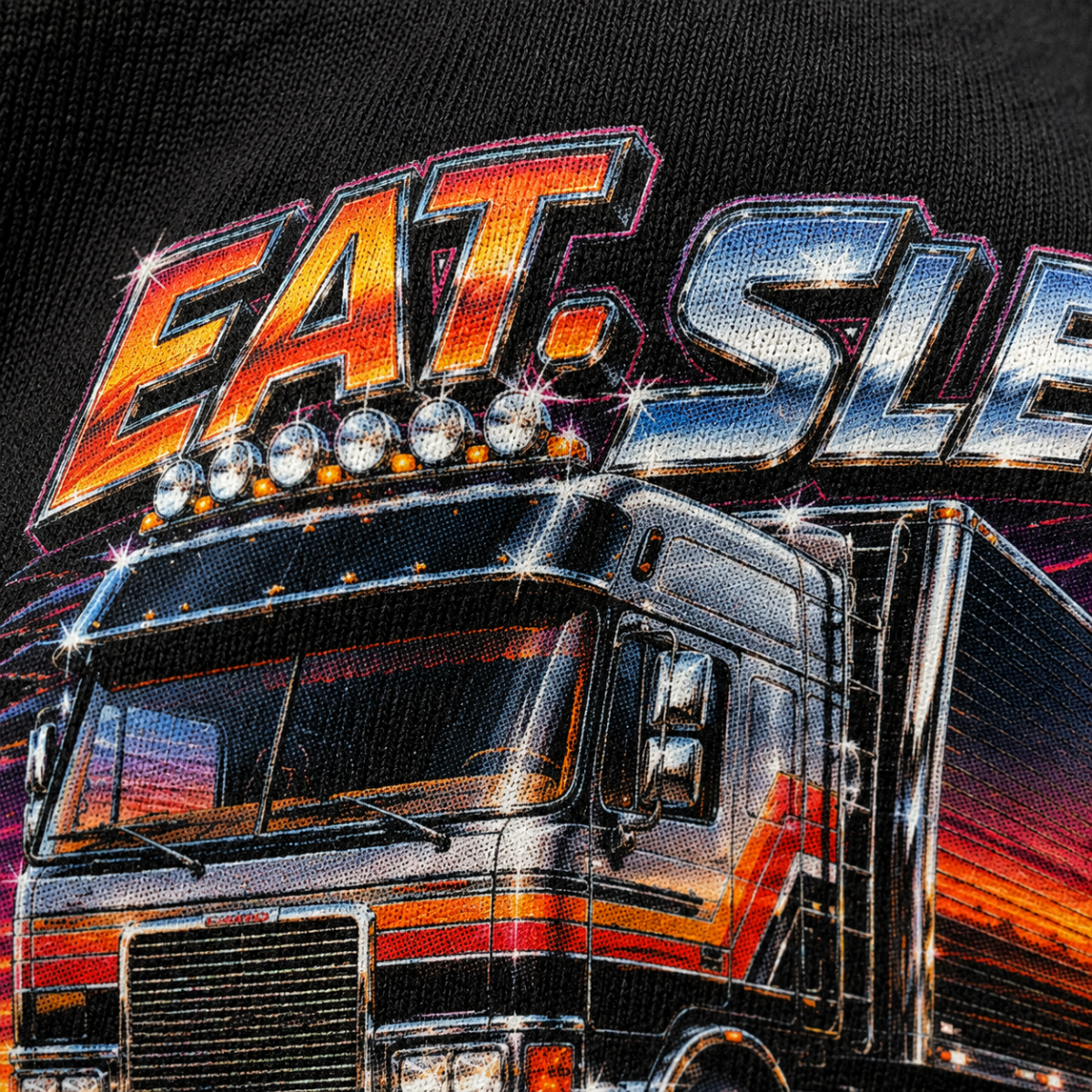 Close-up of a black graphic T-shirt featuring a large stylized truck and the words “EAT SLEEP” in bold metallic lettering with orange, red, and silver highlights.