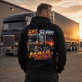 A man stands with his back to the camera wearing a black hoodie with a large graphic print on the back featuring a semi-truck and the text 'EAT SLEEP Haul Repeat.' A large semi-truck is parked in the background at sunset.