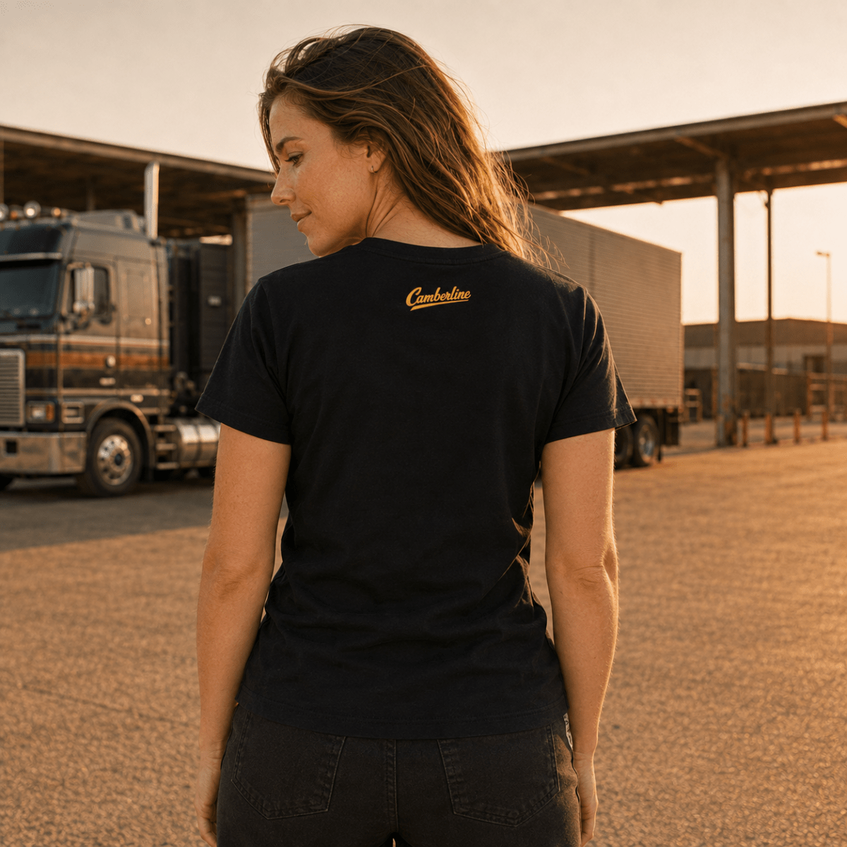 A woman stands outdoors in a truck yard at sunset wearing a black short-sleeve T-shirt with a small yellow script logo on the upper back. A semi-truck and industrial buildings are visible in the background.