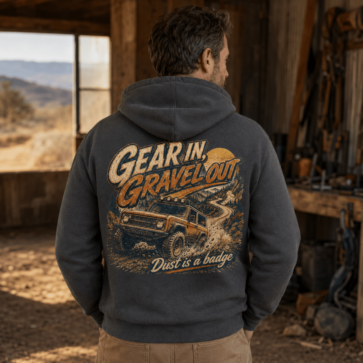 A man viewed from behind wearing a dark gray hoodie with a large back graphic that reads 'Gear in, Gravel out' and 'Dust is a badge,' featuring an off-road vehicle illustration. He is standing inside a rustic workshop or shed with outdoor scenery visible through a window.