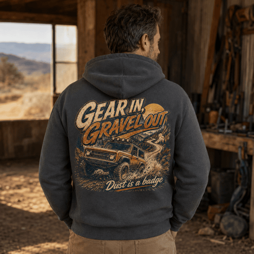 A man viewed from behind wearing a dark gray hoodie with a large back graphic that reads 'Gear in, Gravel out' and 'Dust is a badge,' featuring an off-road vehicle illustration. He is standing inside a rustic workshop or shed with outdoor scenery visible through a window.