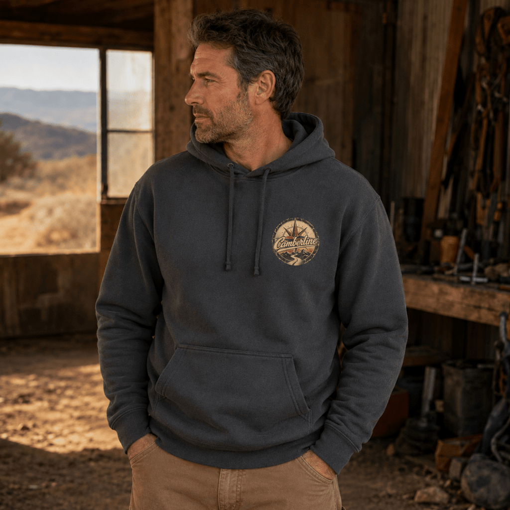 A man wearing a charcoal gray pullover hoodie with a circular embroidered logo on the left chest, standing in a rustic workshop with a mountain landscape visible through a window behind him.