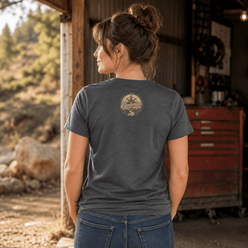 A woman viewed from behind wearing a dark charcoal gray t-shirt with a circular graphic on the upper back, standing in a rustic garage or workshop environment.