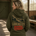 A woman wearing an olive green hoodie with a large graphic of a Lancaster bomber plane and the text 'LANCASTER BOMBER COMMAND' on the back, standing inside a rustic wooden hangar.