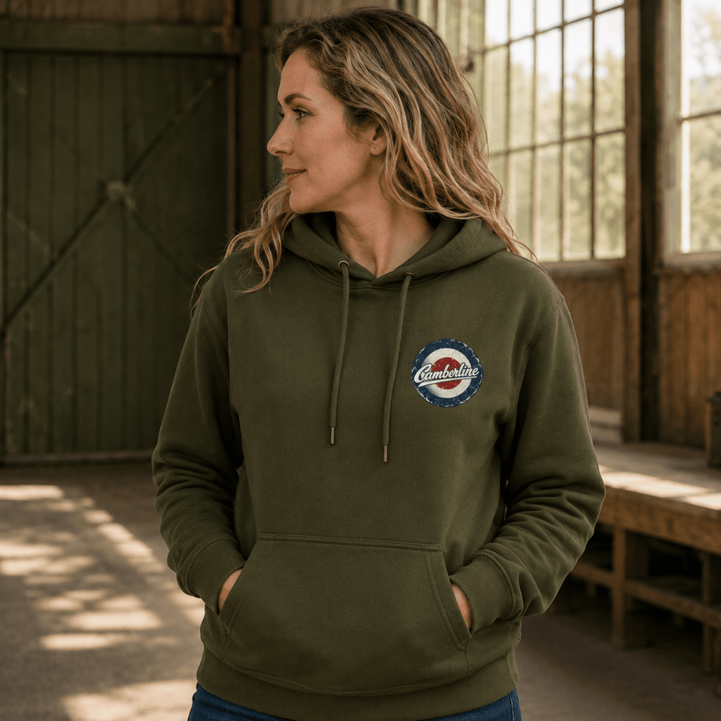 A woman wearing an olive green pullover hoodie with a circular 'Comfort Colors' logo patch on the left chest, standing in a rustic barn interior.