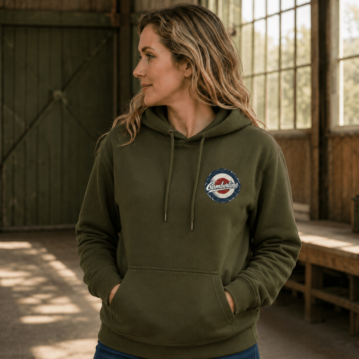 A woman wearing an olive green pullover hoodie with a circular 'Comfort Colors' logo patch on the left chest, standing in a rustic barn interior.