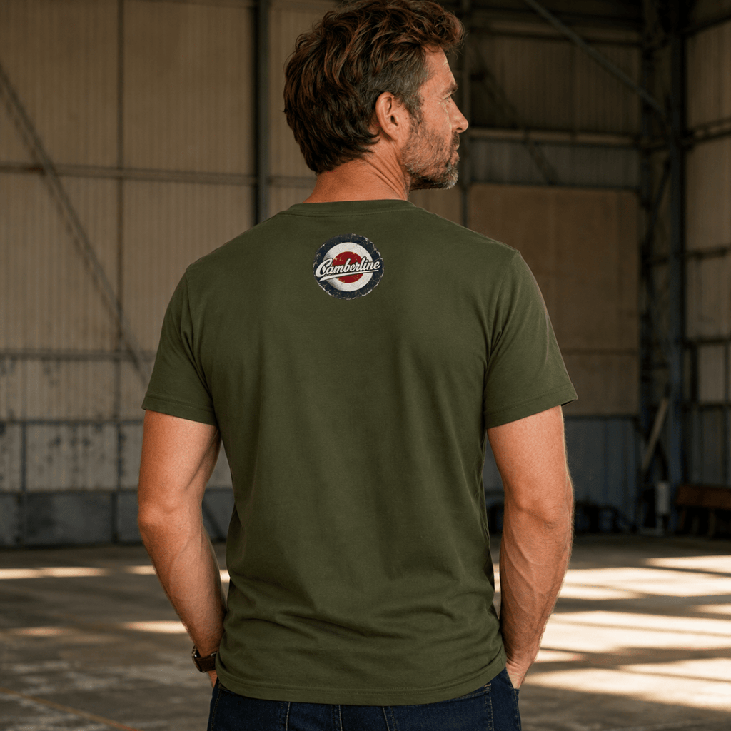A man wearing an olive green t-shirt with a circular 'Camelot' logo on the back, standing inside a large industrial warehouse.