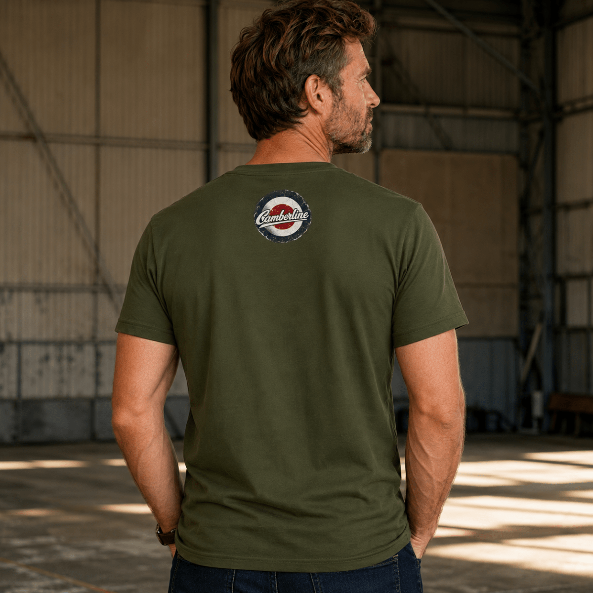 A man wearing an olive green t-shirt with a circular 'Camelot' logo on the back, standing inside a large industrial warehouse.