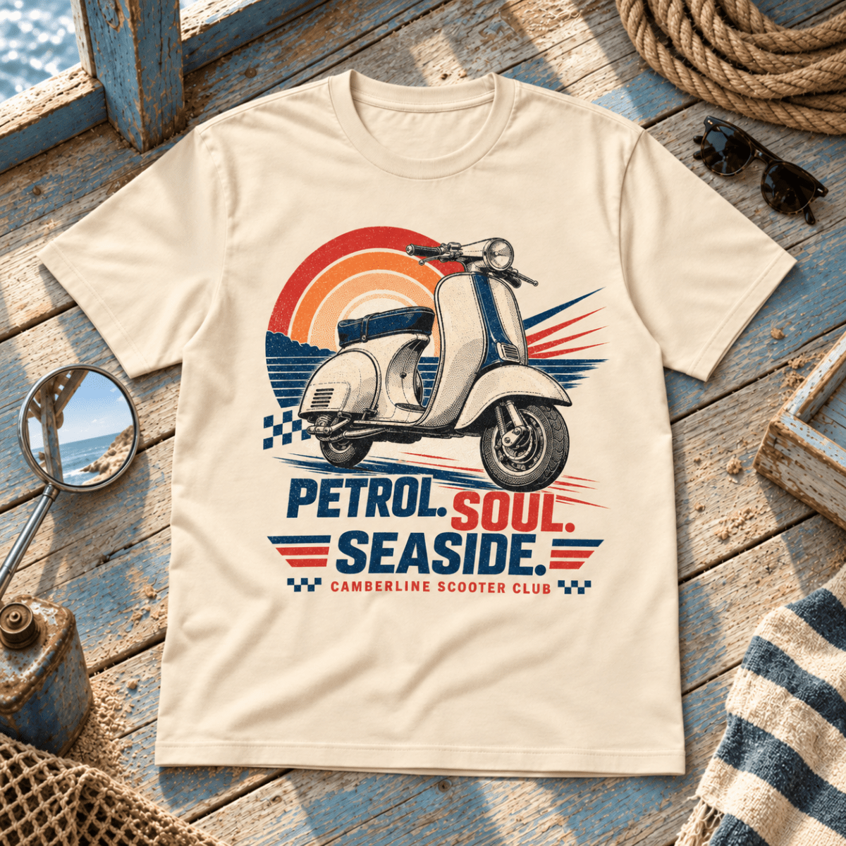 A cream-colored short-sleeve t-shirt with a retro scooter graphic and the text 'PETROL SOUL SEASIDE CAMBERLINE SCOOTER CLUB', displayed in a nautical-themed flat-lay setting on a weathered blue wooden deck with sunglasses, rope, and a striped towel.