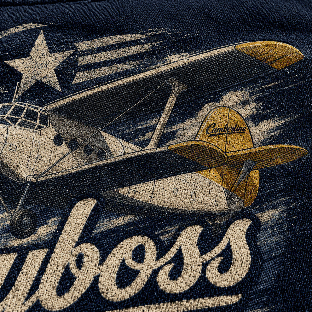 Close-up of a vintage-style graphic print on dark navy fabric, featuring a distressed cream airplane with a yellow tail fin, a large cream star, and bold cream script text reading 'uboss'.