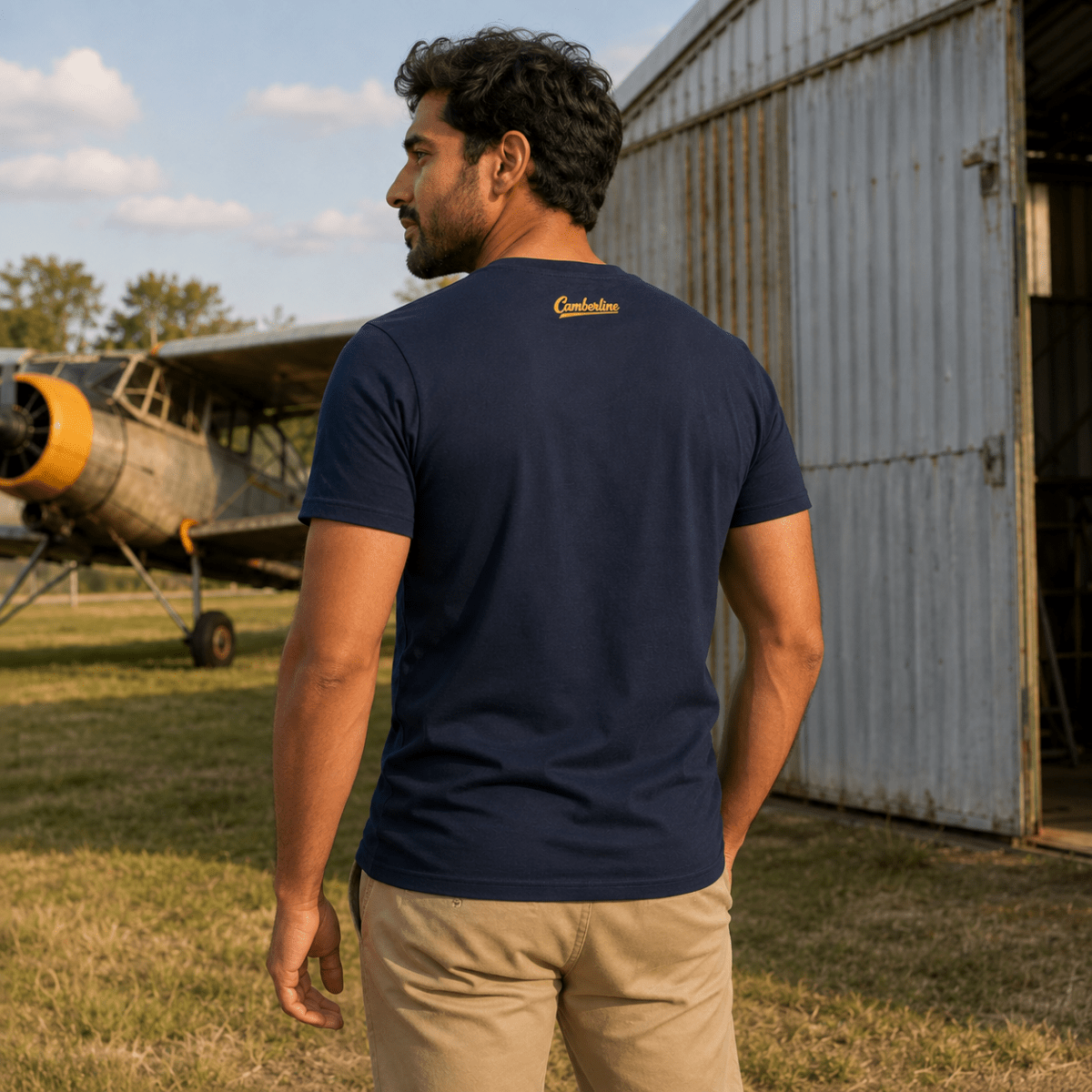 A man wearing a navy blue t-shirt with a gold embroidered 'Cruel Wolf' logo on the back, standing in front of a rustic metal hangar and an old airplane.