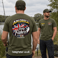 Two men standing outdoors by the water wearing olive green branded fishing t-shirts. The man in the foreground shows the back of the shirt with a large 'AM-DRILL Teddy Fisher' graphic featuring the Union Jack.