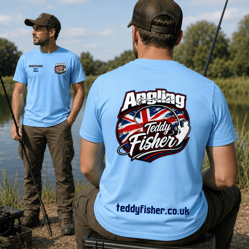 Two men outdoors by a lake wearing light blue fishing t-shirts. The man in the foreground shows the back of the shirt with a large graphic reading 'Angling Teddy Fisher' over a Union Jack design, and the man in the background shows the front of the shirt with a small chest logo and text.