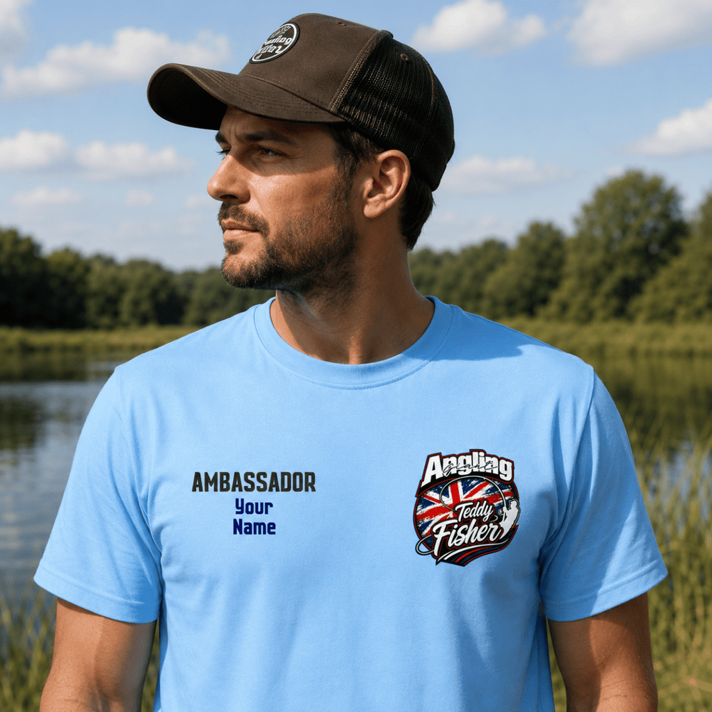 A man wearing a light blue t-shirt and a dark brown trucker hat stands outdoors by a lake. The t-shirt features a custom graphic on the left chest with the text 'AMBASSADOR Your Name' and a 'Angling Today Fisherman' logo on the right chest.