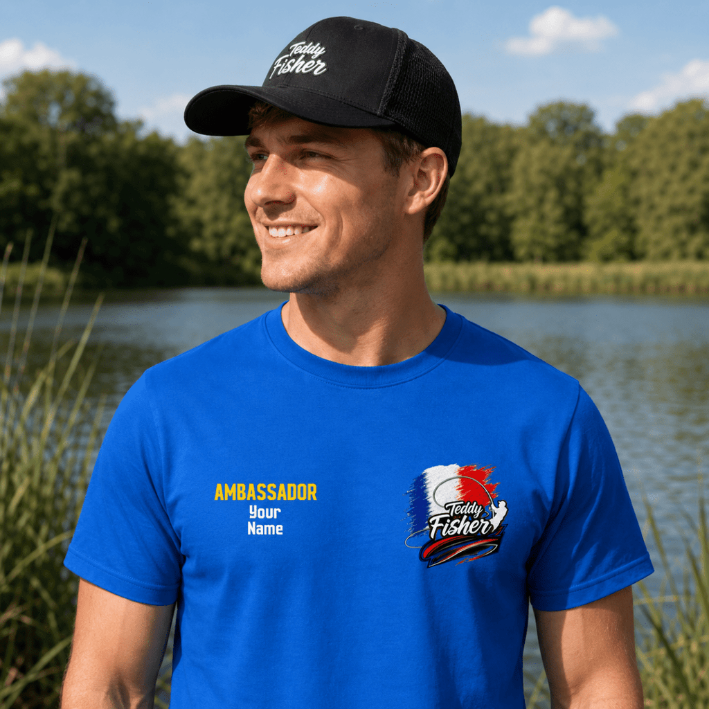 A man wearing a bright blue custom t-shirt and a black trucker hat stands outdoors by a lake. The t-shirt features a custom graphic on the left chest with the text 'AMBASSADOR Your Name' and a custom logo on the right chest.