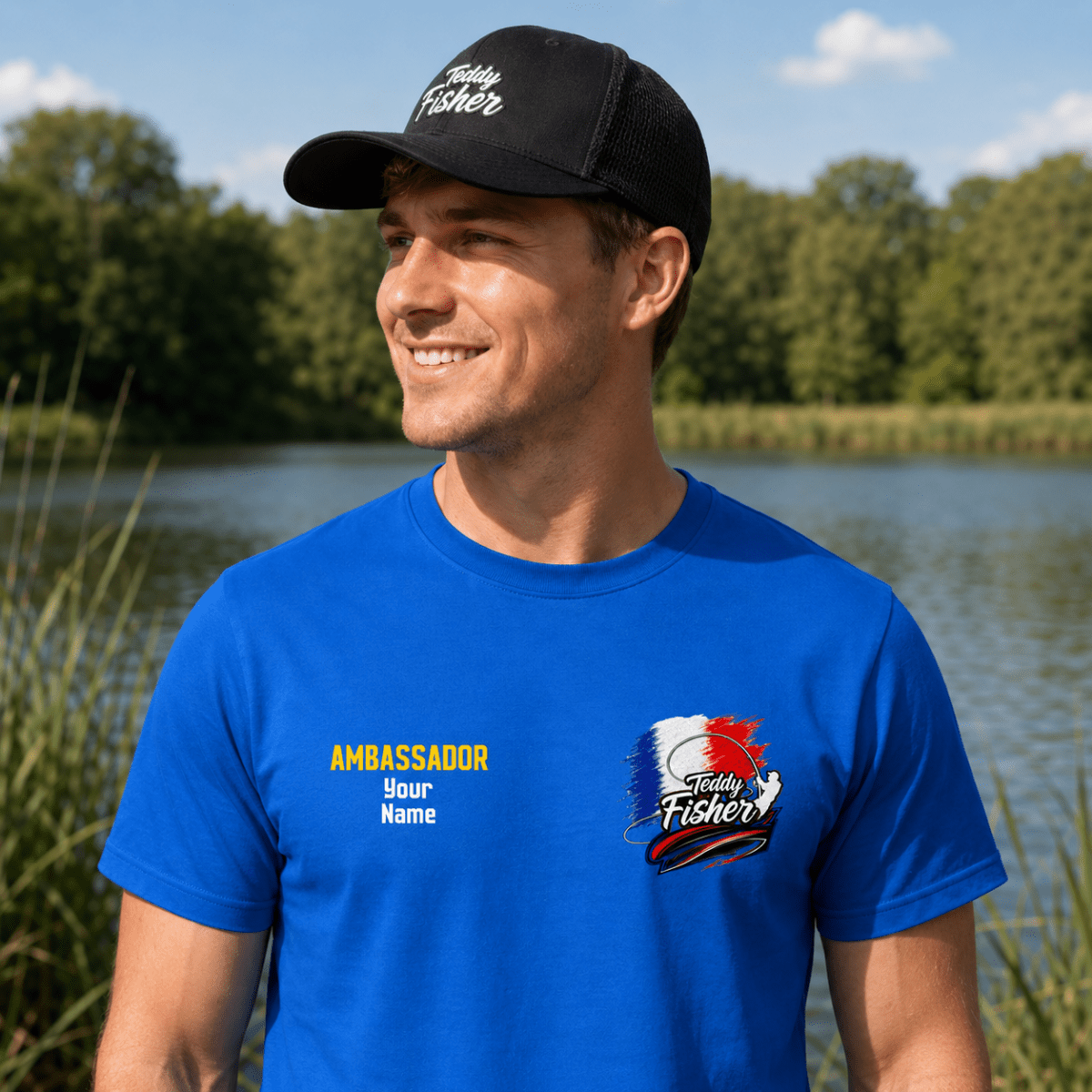 A man wearing a bright blue custom t-shirt and a black trucker hat stands outdoors by a lake. The t-shirt features a custom graphic on the left chest with the text 'AMBASSADOR Your Name' and a custom logo on the right chest.