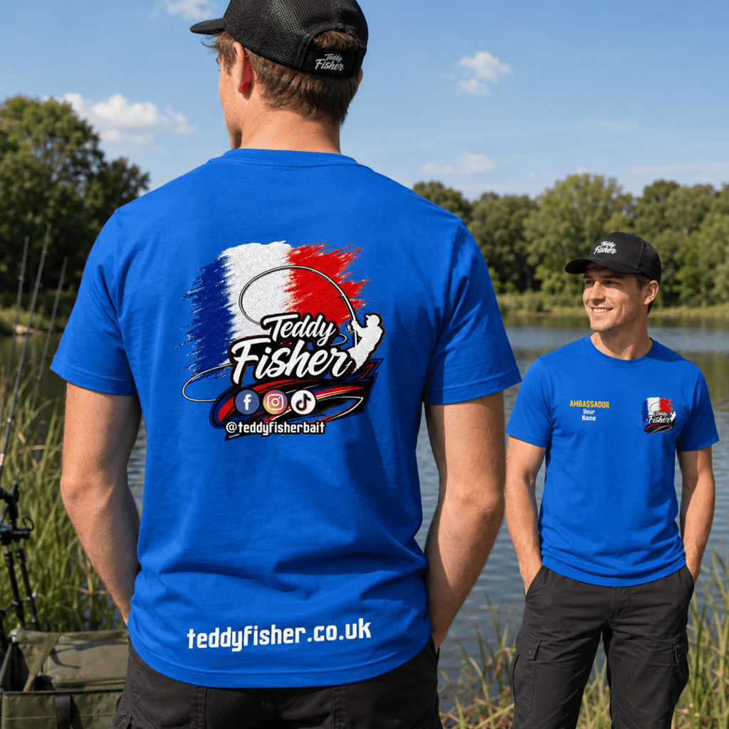 Two men wearing bright blue t-shirts outdoors by a lake. The man in the foreground shows the back of the shirt with a large 'Teddy Fisher' graphic in red, white, and blue, social media icons, and 'teddyfisher.