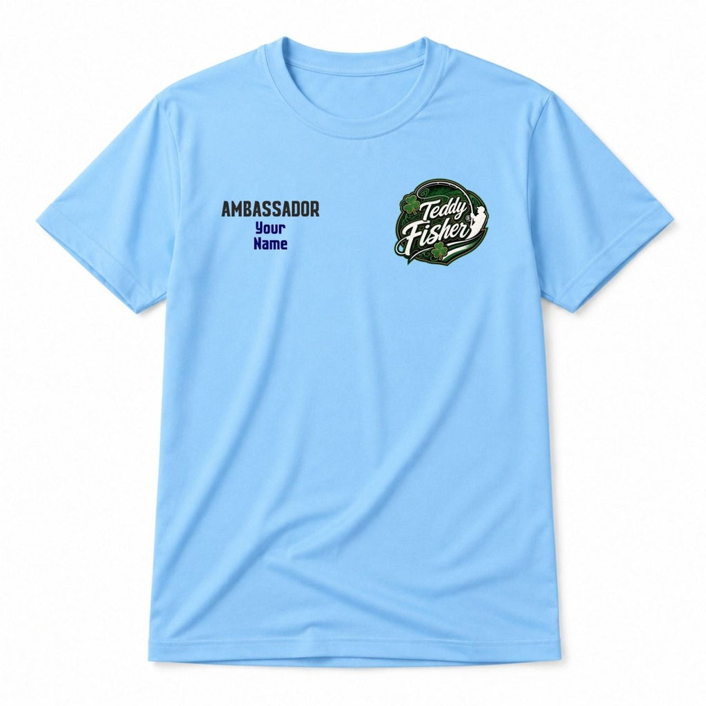 A light blue short-sleeve t-shirt with custom printed graphics on the chest. The left chest features the text 'AMBASSADOR Your Name' in dark blue, and the right chest features a circular green and white graphic reading 'Teddy Fisher'.