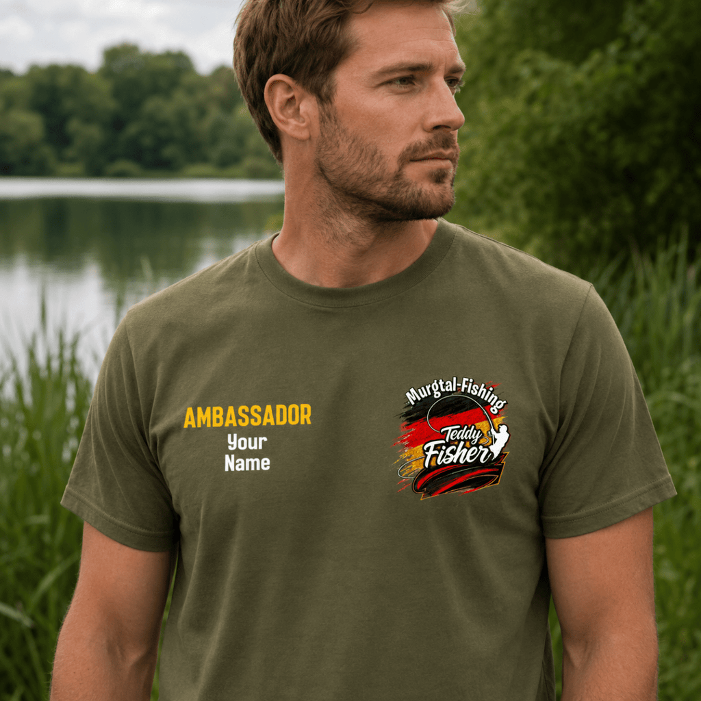 A man wearing an olive green t-shirt with custom printed graphics, standing outdoors in front of a lake and reeds. The t-shirt features a yellow 'AMBASSADOR Your Name' print on the right chest and a 'Murgai Fishing Fisherman' graphic on the left chest.