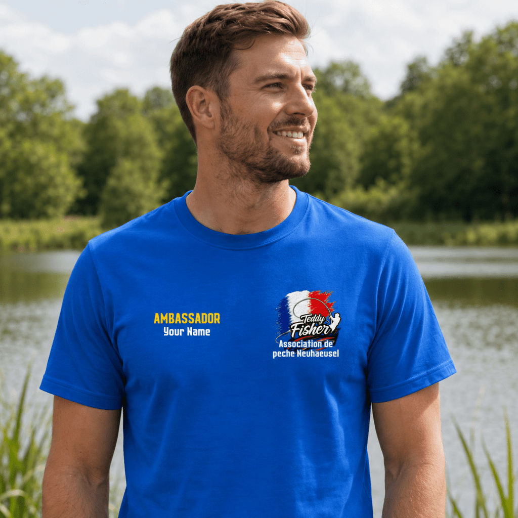 A man wearing a bright blue custom printed t-shirt with a fishing-themed graphic and text, standing outdoors in front of a lake and trees.