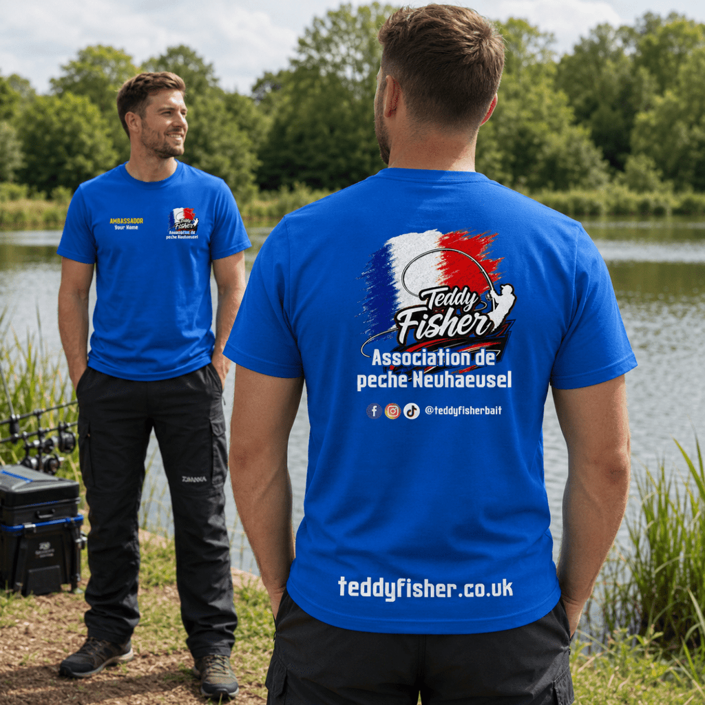 Two men standing by a lake wearing matching royal blue t-shirts. The man in the foreground shows the back of the shirt with a large 'Teddy Fisher Association de peche Neuhaeusel' graphic, while the man in the background shows the front with a small logo.