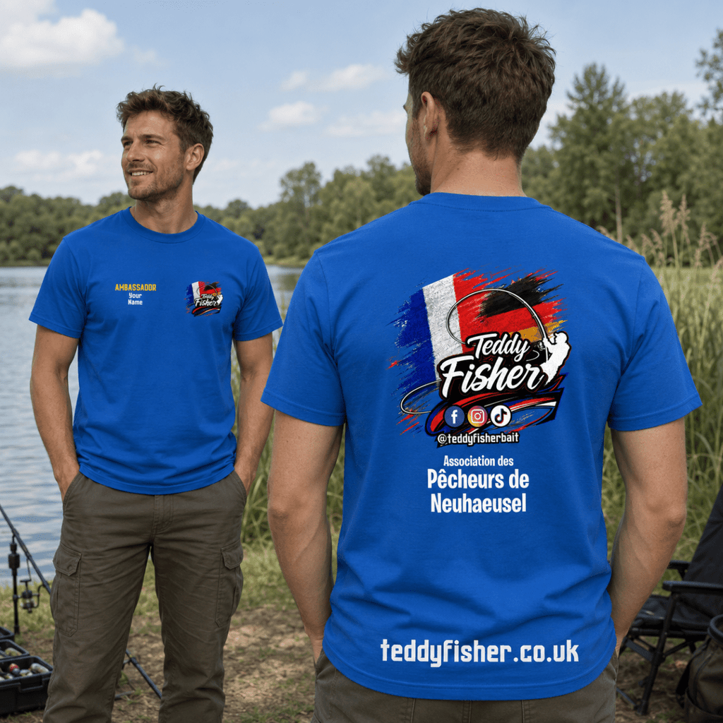 Two men standing outdoors by a lake wearing blue t-shirts. The man on the right shows the back of the shirt with a large 'Teddy Fisher' fishing graphic featuring a French flag motif.