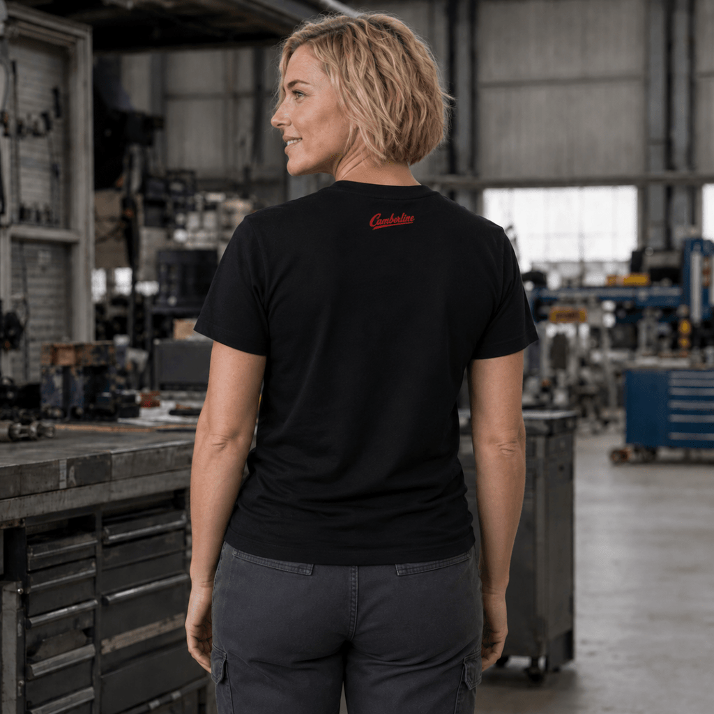 A woman wearing a black short-sleeve t-shirt with a small red script logo on the back, standing in a workshop environment.