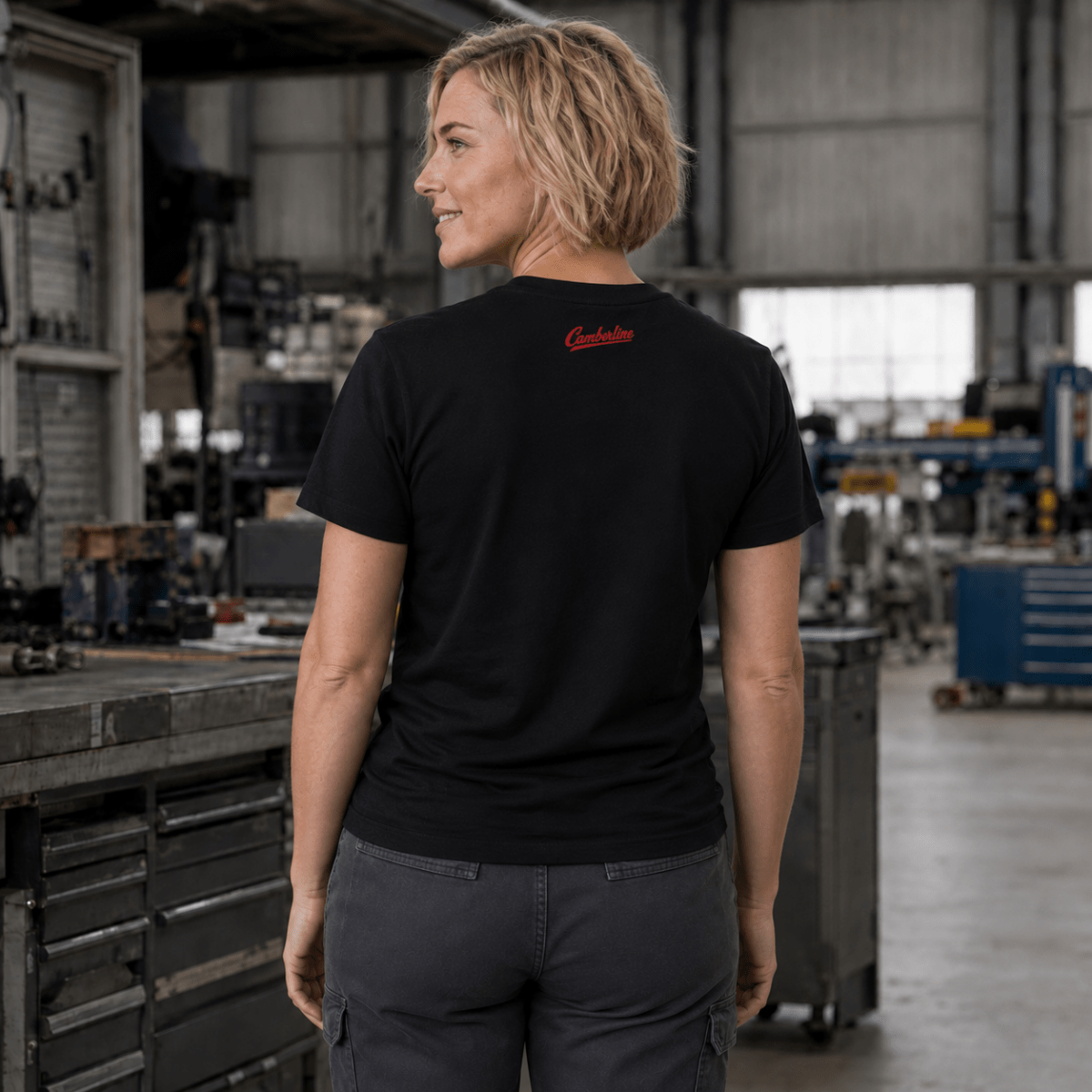 A woman wearing a black short-sleeve t-shirt with a small red script logo on the back, standing in a workshop environment.